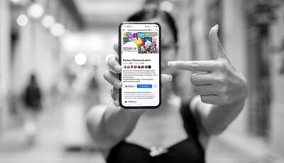 Una donna attraente con un vestito nero mostra uno smartphone dove è visualizzata la pagina Facebook dell'Agenzia Factory Communication