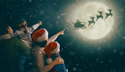 papà, mamma e 2 figli guardano la slitta di babbo natale trainata dalle renne che vola in cielo