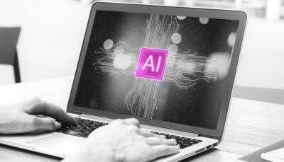 Foto in bianco e nero. Due mani stanno scrivendo su un computer portatile. In centro alla schermo, in un riquadro quadrato di colore viola Factory Communication la scritta AI