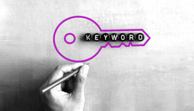 su un foglio grigio è stato disegnato il profilo di una chiave. Nella chiave la parola Keywords scritta sui dadi. In primo piano una mano con una penna. L'immagine è in bianco e nero, solo la chiave ha il profilo di colore viola Factory Communication.
