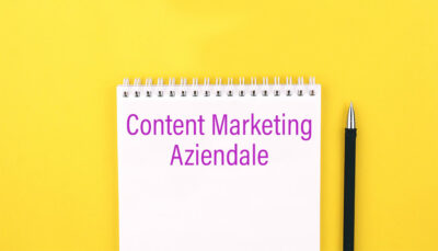 Scritta content marketing aziendale di colore viola su un block notes di colore bianco. Alla destra del block notes una matita. Il colore di sfondo è il giallo factory communication