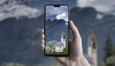 Una mano regge un cellulare sul cui schermo è visibile l'immagine panoramica di una chiesa con alle spalle montagne e alberi, sotto un cielo blu e nuvoloso. Lo sfondo dell'immagine mostra un paesaggio montuoso simile.