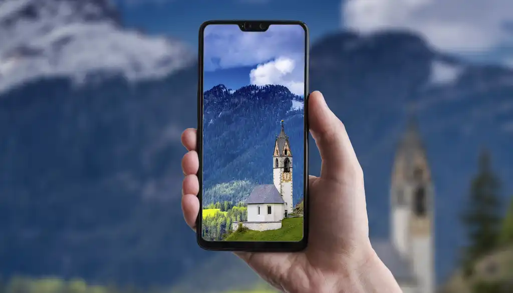 Una mano regge un cellulare sul cui schermo è visibile l'immagine panoramica di una chiesa con alle spalle montagne e alberi, sotto un cielo blu e nuvoloso. Lo sfondo dell'immagine mostra un paesaggio montuoso simile.